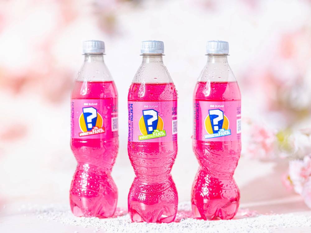 Tre rosa Fanta-flasker med spørsmålstegn på etiketten står på en lys overflate med en rosa og hvit bakgrunn.