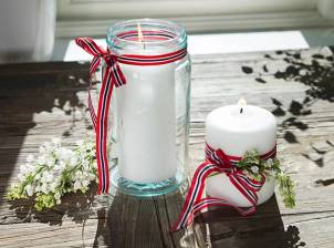 To lyslykter dekorert med røde, hvite og blå bånd, omgitt av blomster, på en utendørs trebord til 17. mai-feiring.