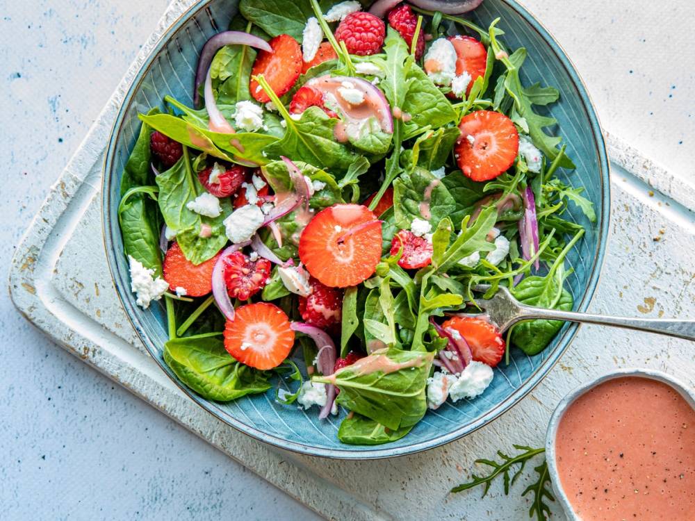 En frisk salat med jordbær, bringebær, chevreost og grønne blader i en skål, med en liten bolle med dressing ved siden av.