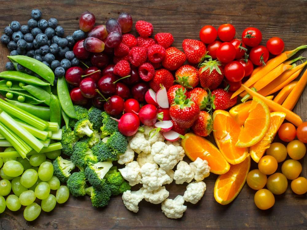 Et utvalg av frukt og grønnsaker, inkludert blåbær, druer, kirsebær, jordbær, cherrytomater, gul paprika, brokkoli, blomkål, og grønne druer, lagt ut på et bord.