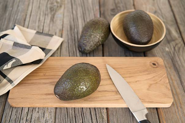 To avokadoer på et skjærebrett med en kniv, klar til å skrelles, og en avokado i en skål i bakgrunnen.