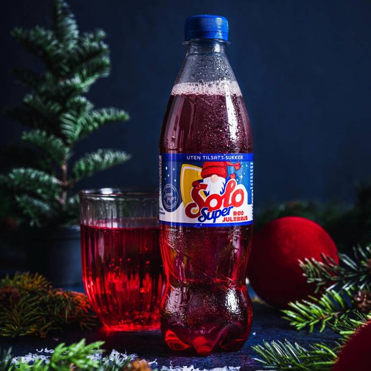 En flaske julebrus med en glass ved siden av, omgitt av julegrener og en rød julekule, på en mørk bakgrunn.