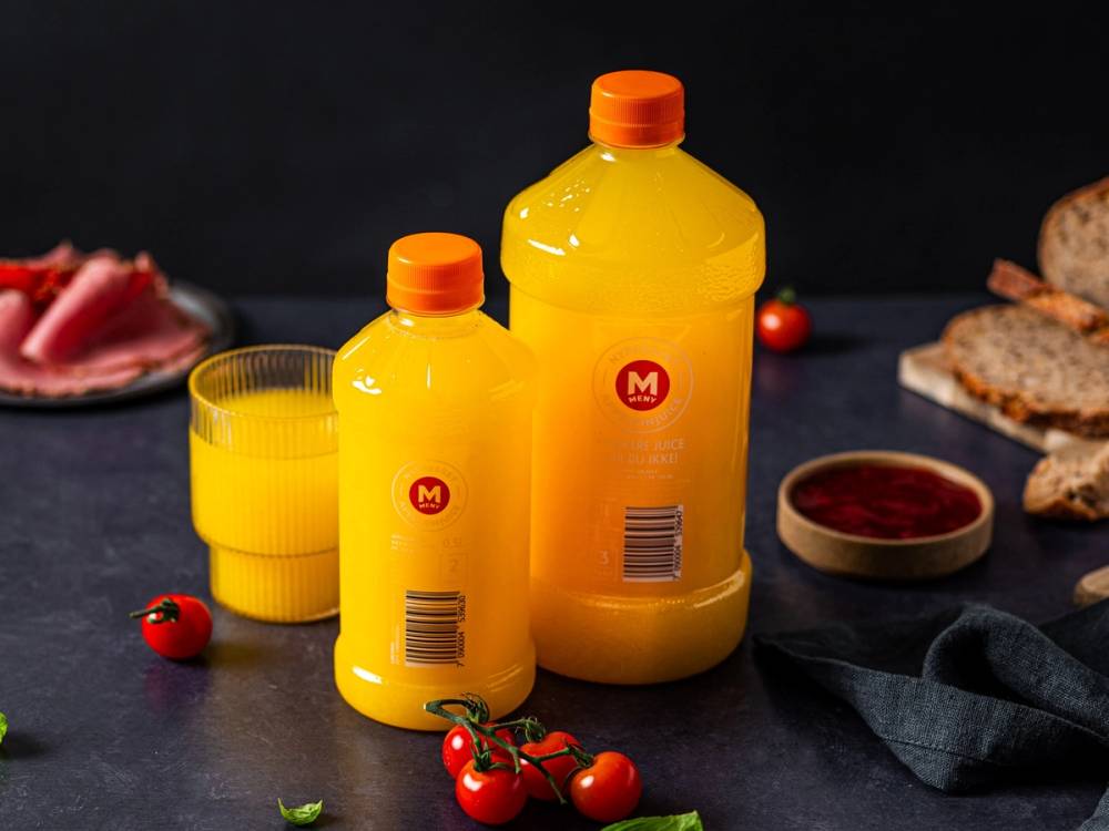 To storegule plastflasker med oransje korker, en liten og en stor, står på et mørkt bord med brød, tomatskiver og en glass med appelsinjuice rundt seg.