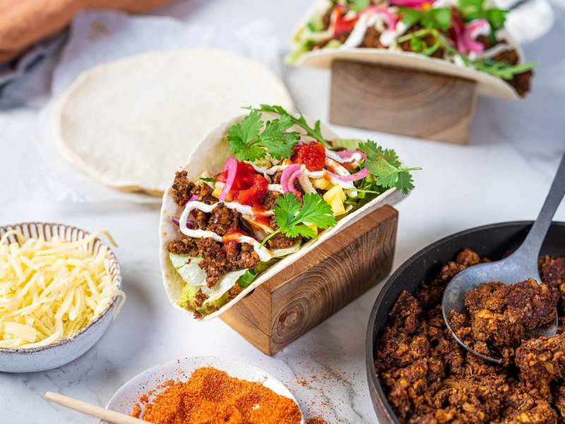 Vegetar taco fylt med grønnsaker, krydret deig og fersk koriander, omgitt av ulike taco-ingredienser og tilbehør.