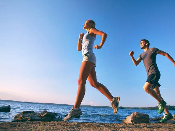 To personer løper langs en strand med vann og steiner, under en klar blå himmel.