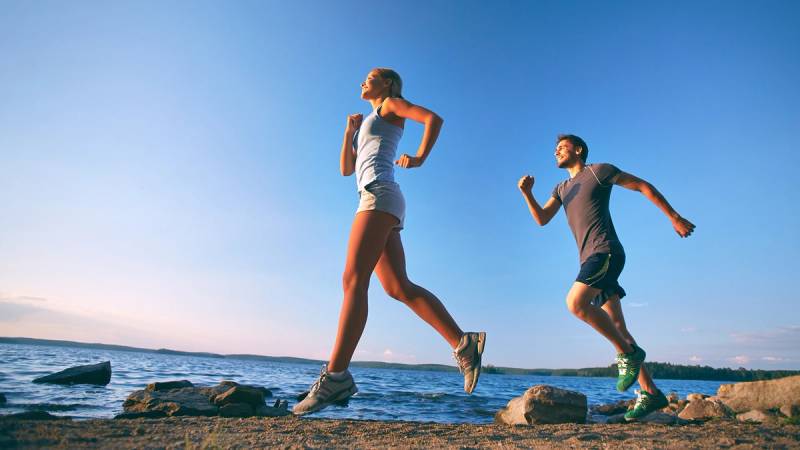 To personer løper langs en strand med vann og steiner, under en klar blå himmel.