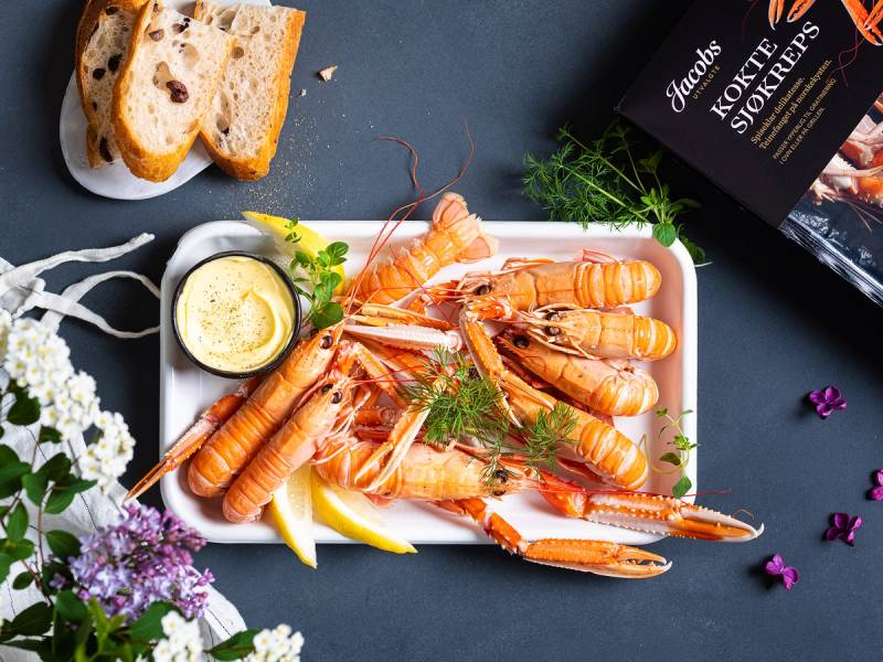 Sjøkreps på en tallerken med sitron, brød og urter, med en pakke Jacobs utvalgte sjøkreps i bakgrunnen