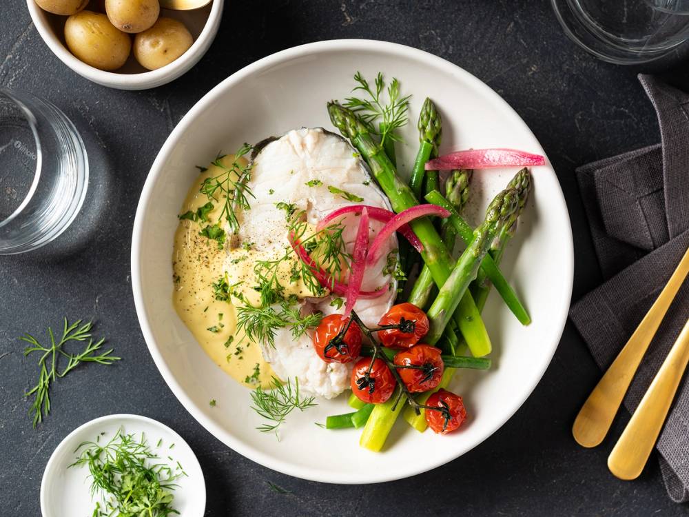 Skrei med hollandaise saus og grønnsaker på en hvit tallerken, med glass vann og bestikk ved siden av.