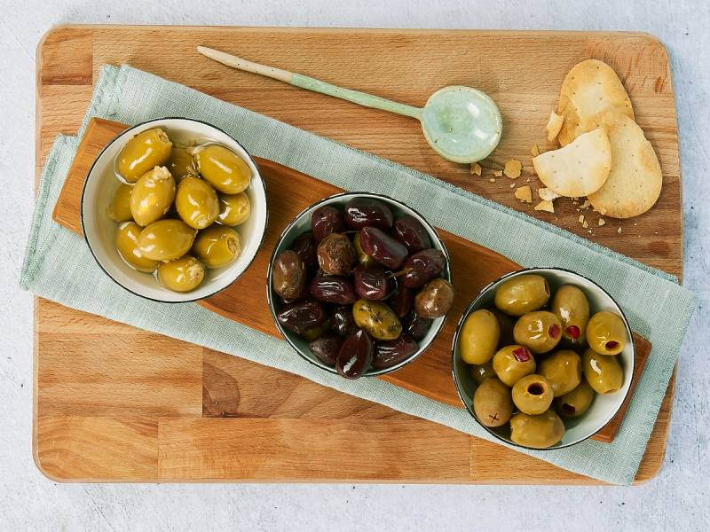 Tre skåler med grønne og svarte oliven på en treplate, med en liten skje og et stykke brød ved siden av.