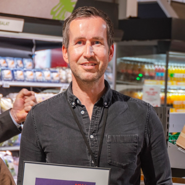 En mann med kort, grått hår i en butikk, holder et sertifikat eller en plakat, med hyller av varer i bakgrunnen.