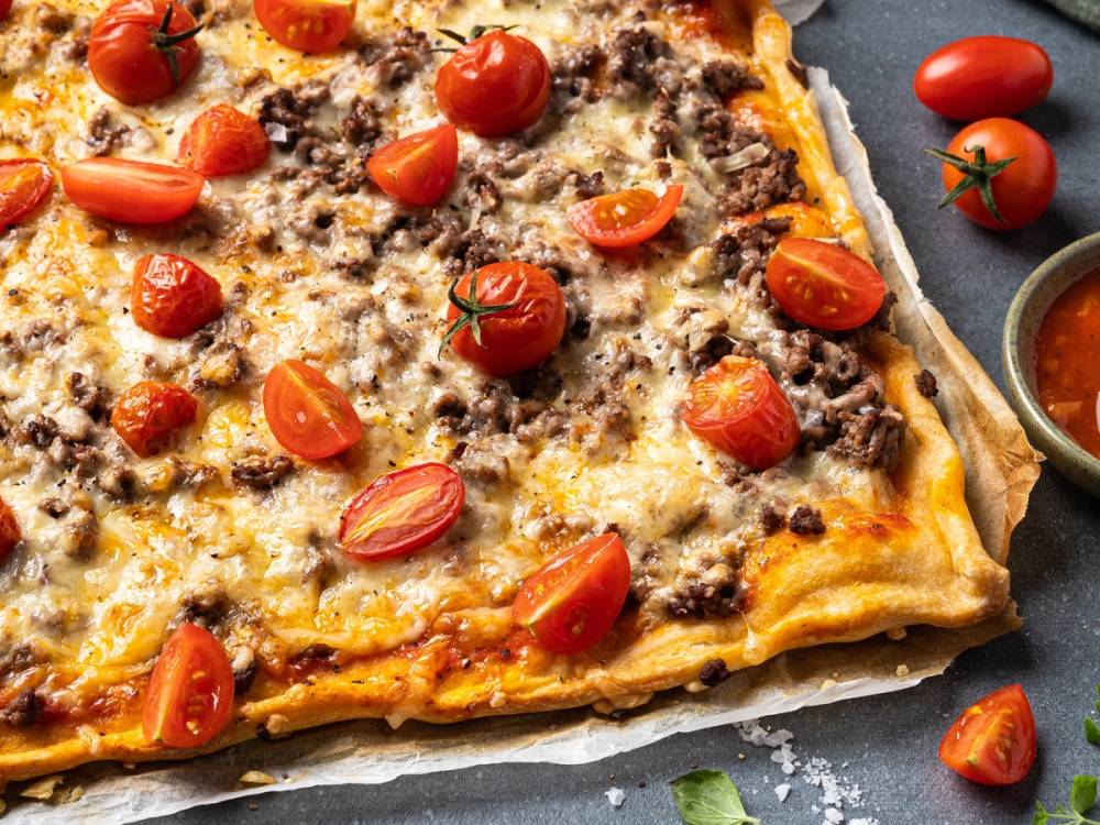 En pizza med kjøttdeig, cherrytomater og ost på en stekeplate med noen tomater og en liten skål med saus rundt.