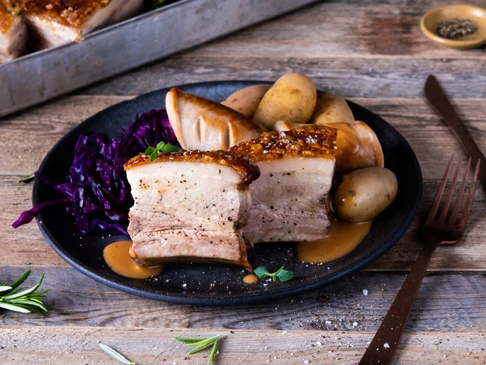 Stekt ribbe med sprø svor, kokte poteter og rødkål på en svart tallerken, plassert på et rustikt trebord.