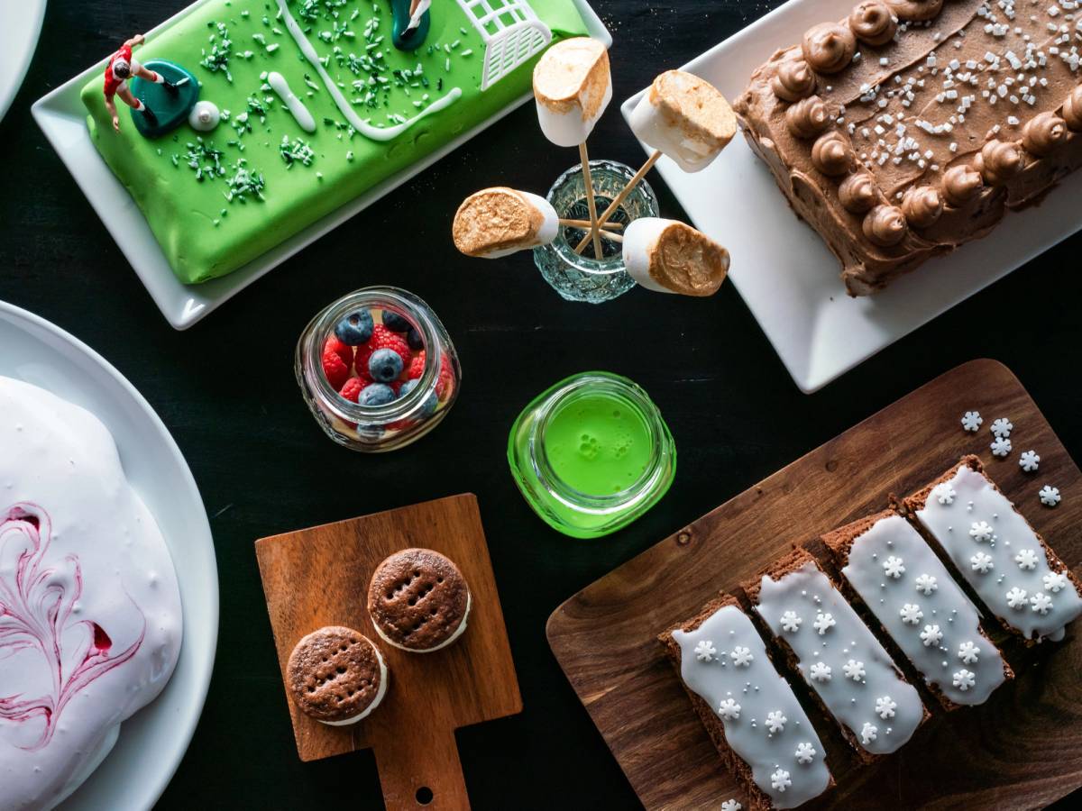 Et utvalg av ulike kaker og småkaker på et bord, inkludert en grønn kake, en sjokoladekake, og småkaker med hvit glasur og snøfnugg, samt en glass med bær og en liten skål med grønn gelé.