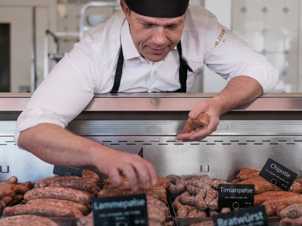 En person i hvit skjorte og svart forkle som selger pølser i en matbod, med ulike typer pølser utstilt foran seg.