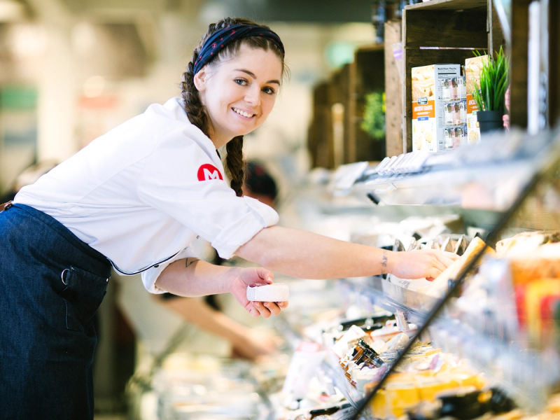En ansatt fyller på ferskvarer i en butikkhyller, med fokus på utvalget av matvarer.