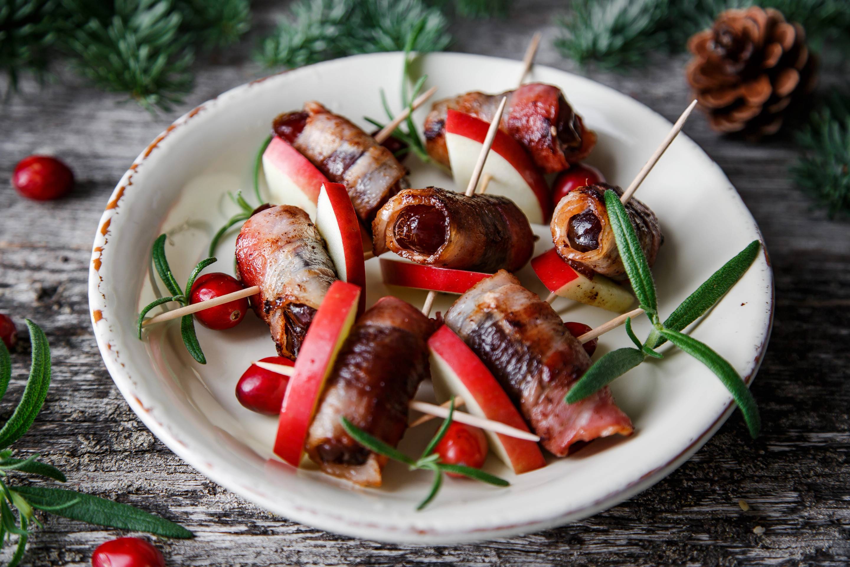 Daddelbacon-ruller servert på en hvit tallerken med urter og små tomater rundt, på et rustikt trebord.