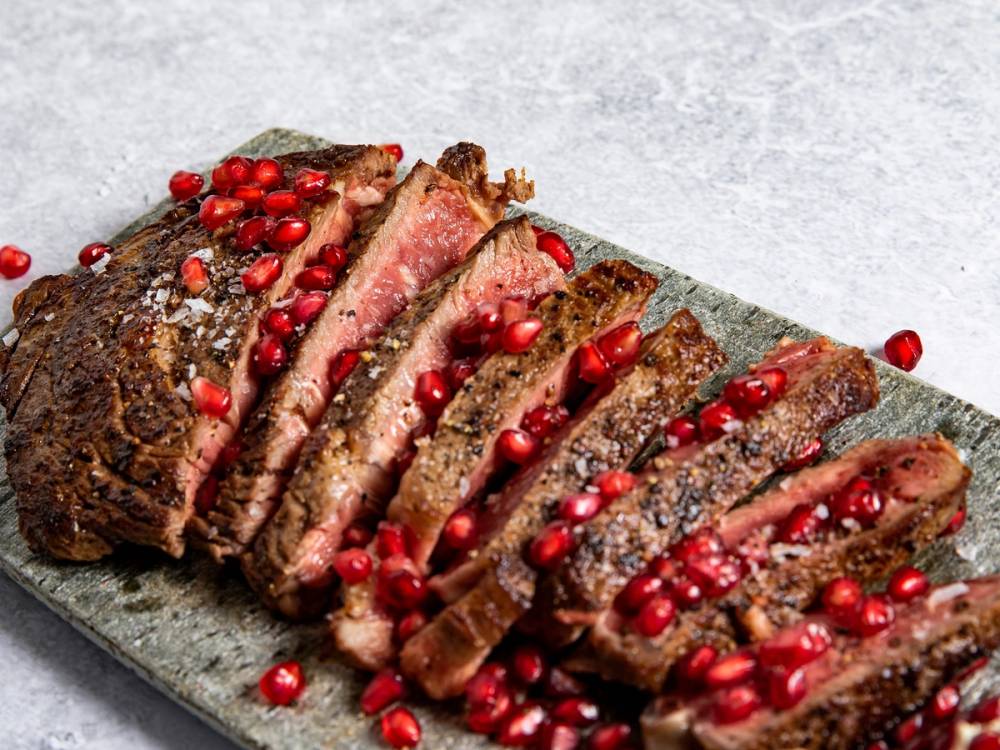 Entrecôte med granateplekjerner skåret i skiver og pyntet med flere granateplekjerner på en skjærefjøl.