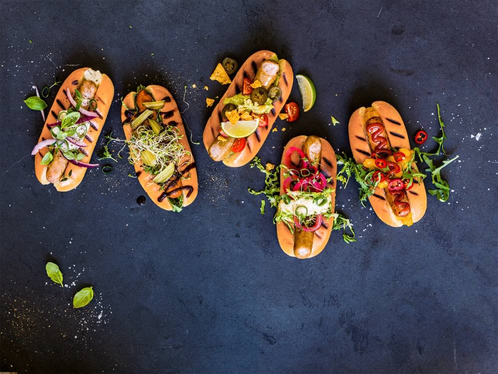 Fem pølser i brød med ulike toppings og grønnsaker på en mørk bakgrunn.