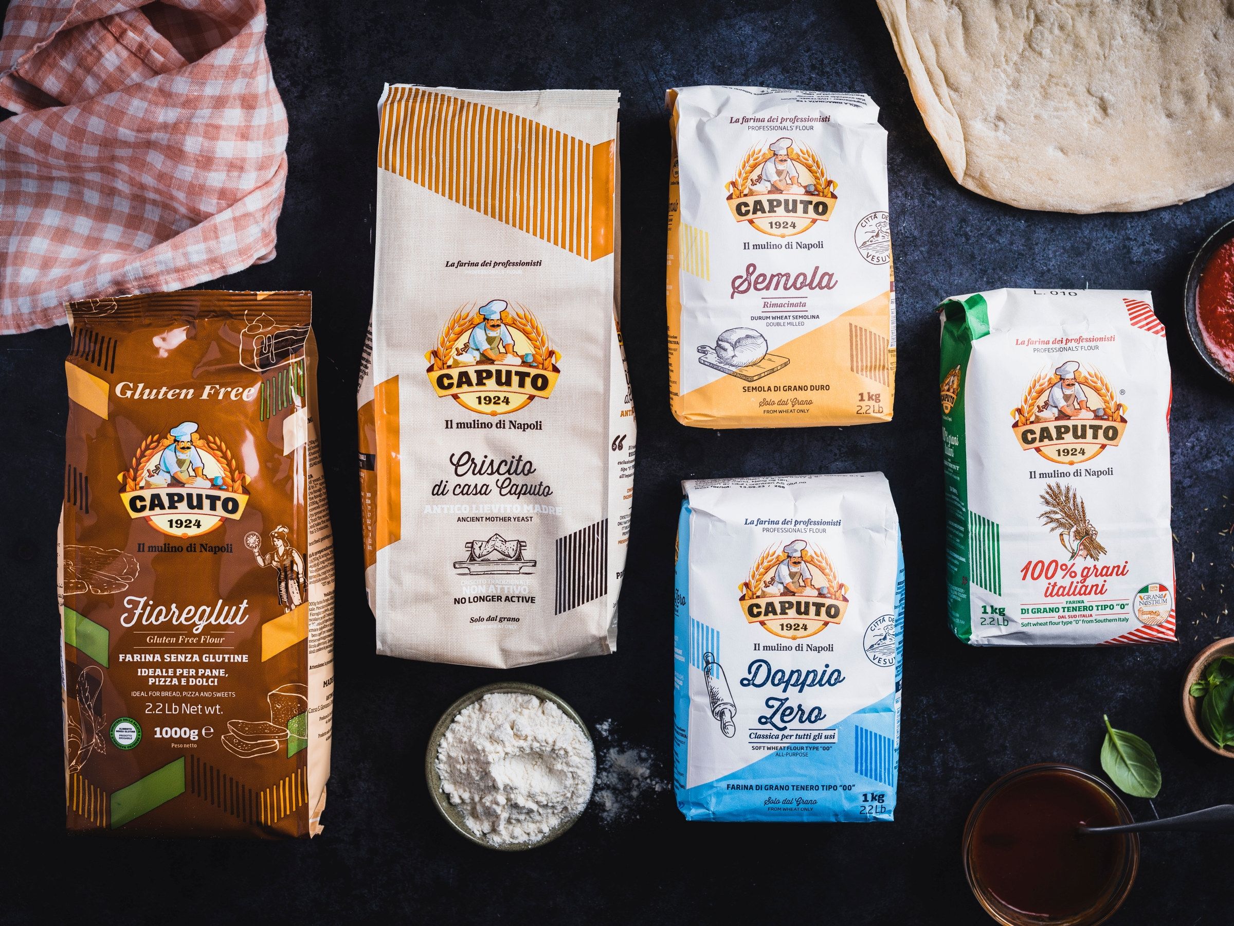 Ulike typer av mel, inkludert hvetemel og fullkornsmel, på et svart bord med noen grønne blader og en liten bolle med mel.