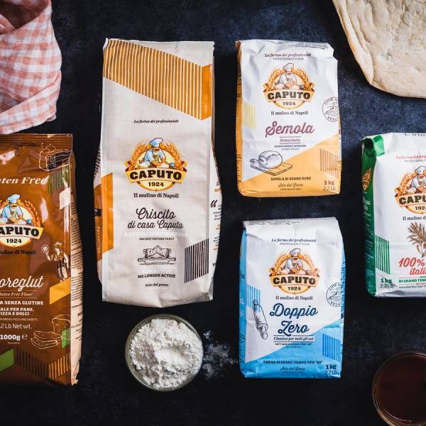 Ulike typer av mel, inkludert hvetemel og fullkornsmel, på et svart bord med noen grønne blader og en liten bolle med mel.