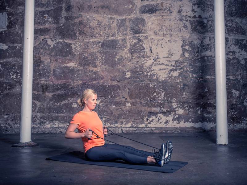 En person sitter på gulvet og bruker en treningsmaskin for roing, med en grå murvegg i bakgrunnen.