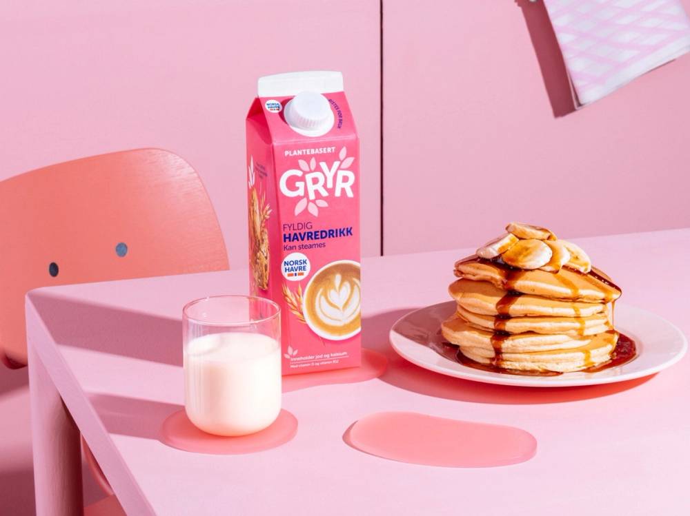 En kopp melk, en pakke med havredrikk og en tallerken med flere pannekaker på et rosa bord med rosa bakgrunn