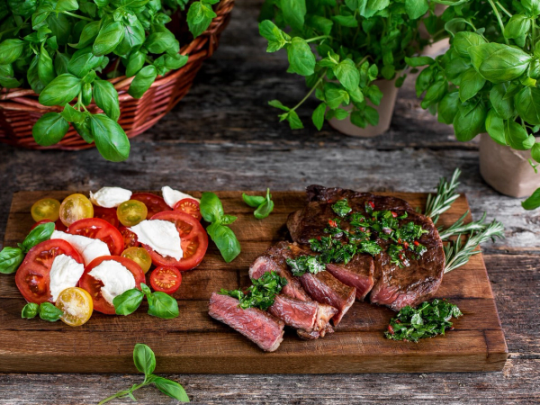 Bilde av en tallerken med biff, urter og tomatsalat med mozzarella og grønne oliven, omgitt av urter i potter og på et trebord.