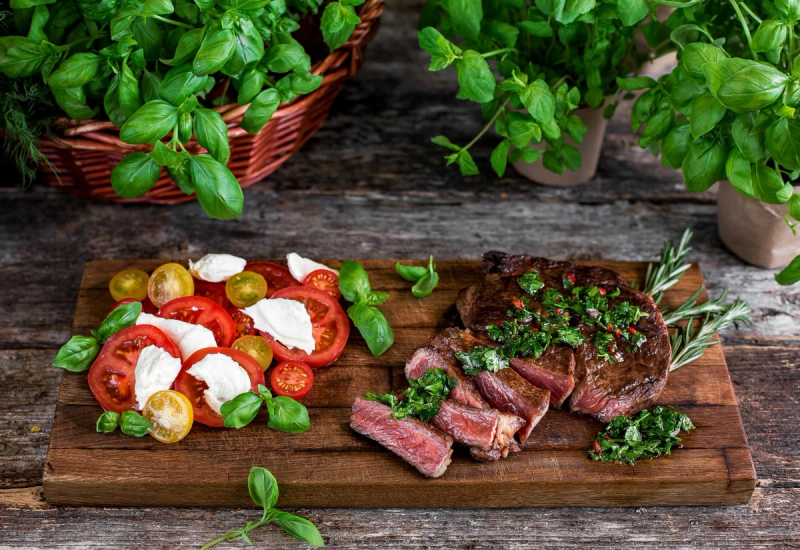 Bilde av en tallerken med biff, urter og tomatsalat med mozzarella og grønne oliven, omgitt av urter i potter og på et trebord.