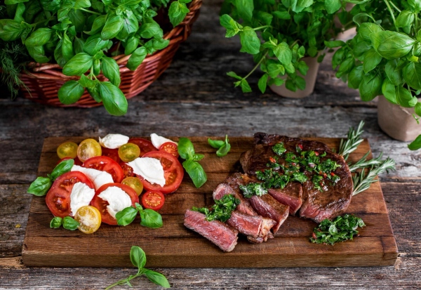 Bilde av en tallerken med biff, urter og tomatsalat med mozzarella og grønne oliven, omgitt av urter i potter og på et trebord.