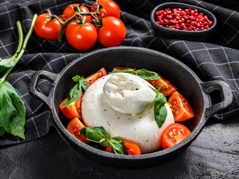 Caprese-salat med tomater, mozzarella og basilikum i en svart skål, omgitt av flere tomater og en liten skål med pepperkorn