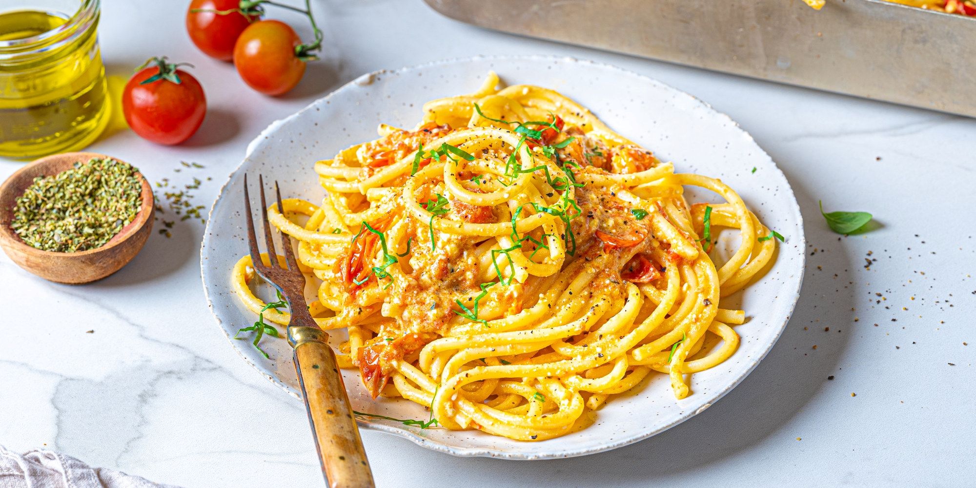 Pasta med fetaost og cherrytomater på en hvit tallerken, omgitt av tomater og krydderurter, klar til servering.