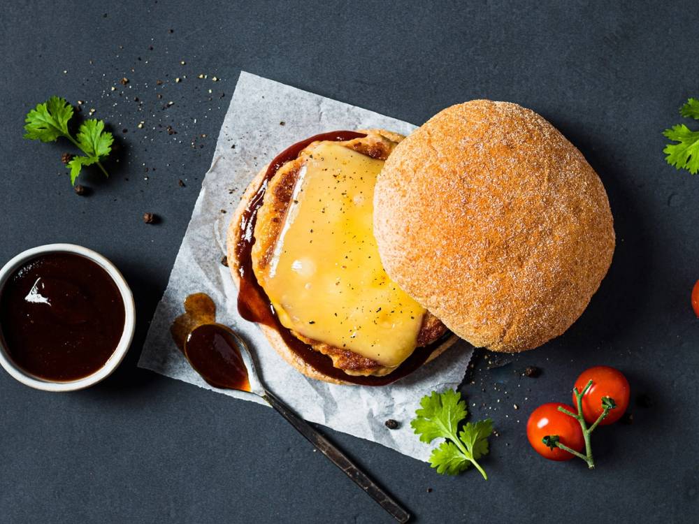 En baconburger med ost og bacon i et brød, servert med små tomater og urter på en mørk bakgrunn.