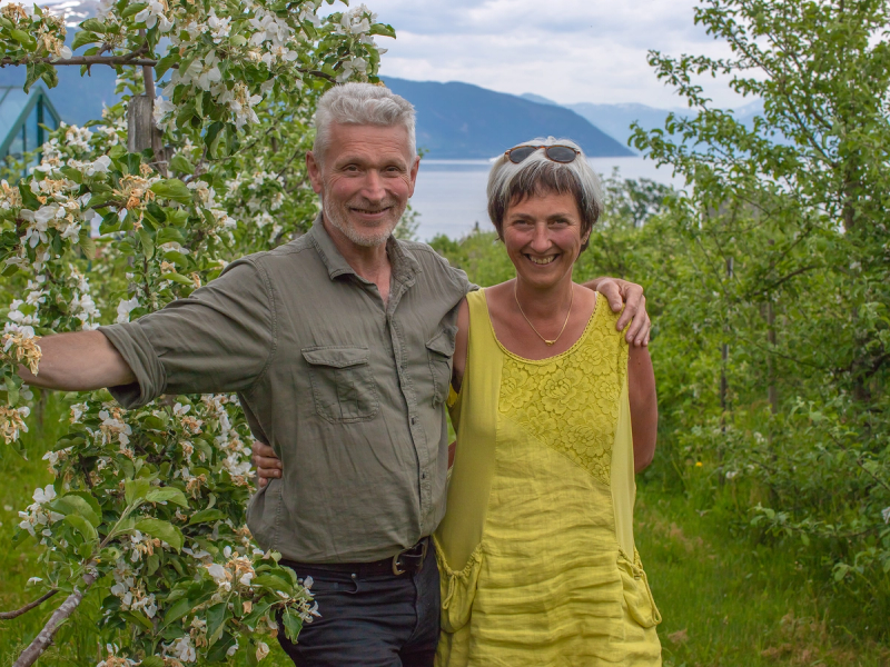 To personer står ute blant blomstrende trær med vann og fjell i bakgrunnen, og de har armene rundt hverandre.