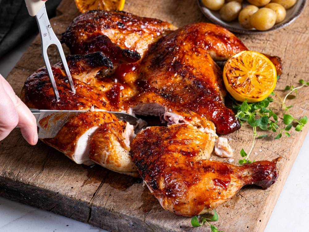Helstekt kylling med sprø skorpe på et skjærebrett, med en halvert sitron og urter ved siden av.