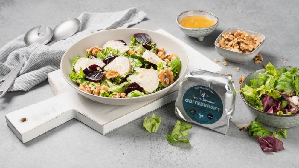 En salat med grønnsaker, ost og oliven i en hvit bolle, omgitt av dressing og nøtter på en grå overflate