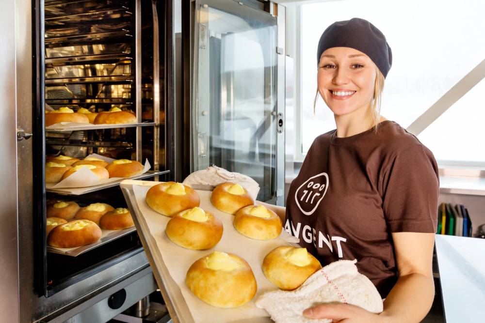 Person med mørk t-skjorte som bærer en brett med nybakte rundstykker ut av en ovn i et bakeri, med en stor ovn i bakgrunnen.