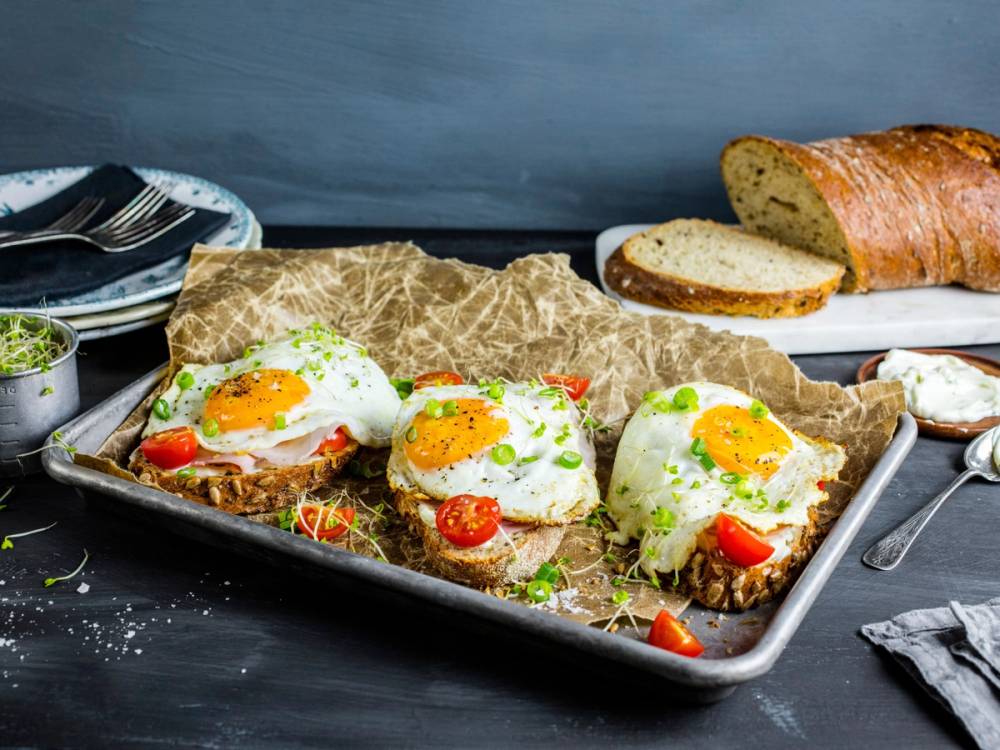 Frokostbord med stekte egg, cherrytomater, brød og en skål med grønt tilbehør.