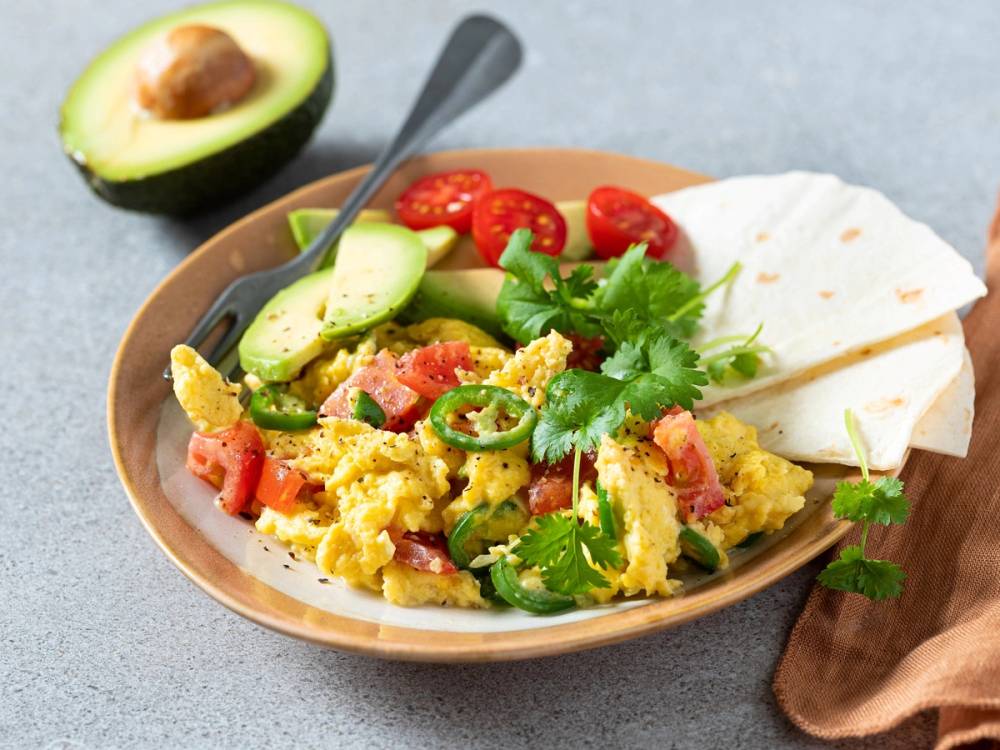 En tallerken med meksikansk eggerøre med grønnsaker, tomater og koriander, sammen med en skive tortilla og en avokado i bakgrunnen.