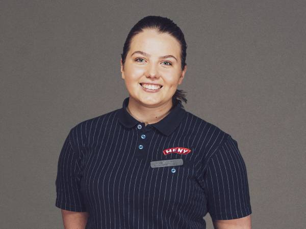 En person i en mørkeblå stripete uniform med navnepin og logo står foran en grå bakgrunn.