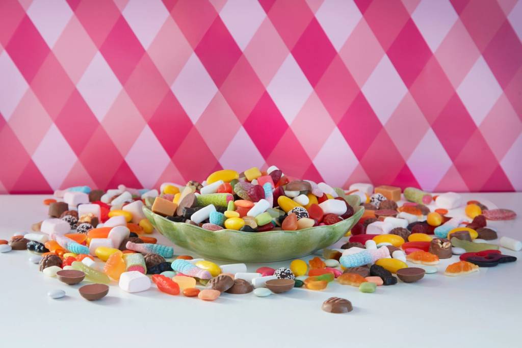 En stor grønn skål fylt med ulike typer godteri og tabletter, plassert på et bord foran en rosa og hvit rutete bakgrunn.