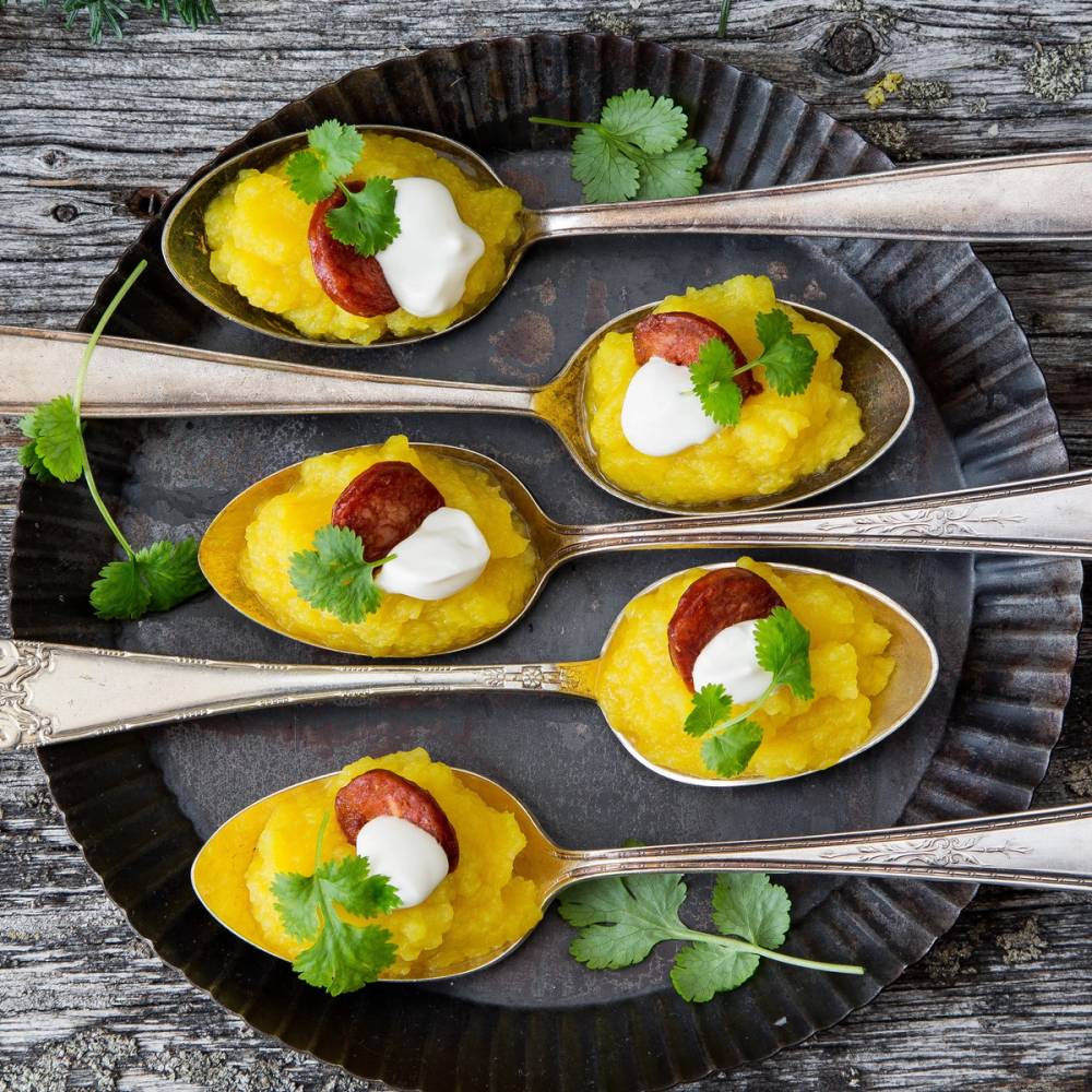 Kålrabi med chorizo servert i små skjeer med urter og en klatt med krem på en svart tallerken, på en treplate.