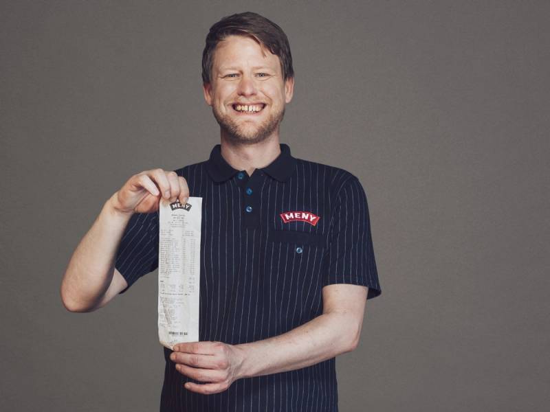 Person som viser en kvittering, med en logo på skjorten, mot en grå bakgrunn.