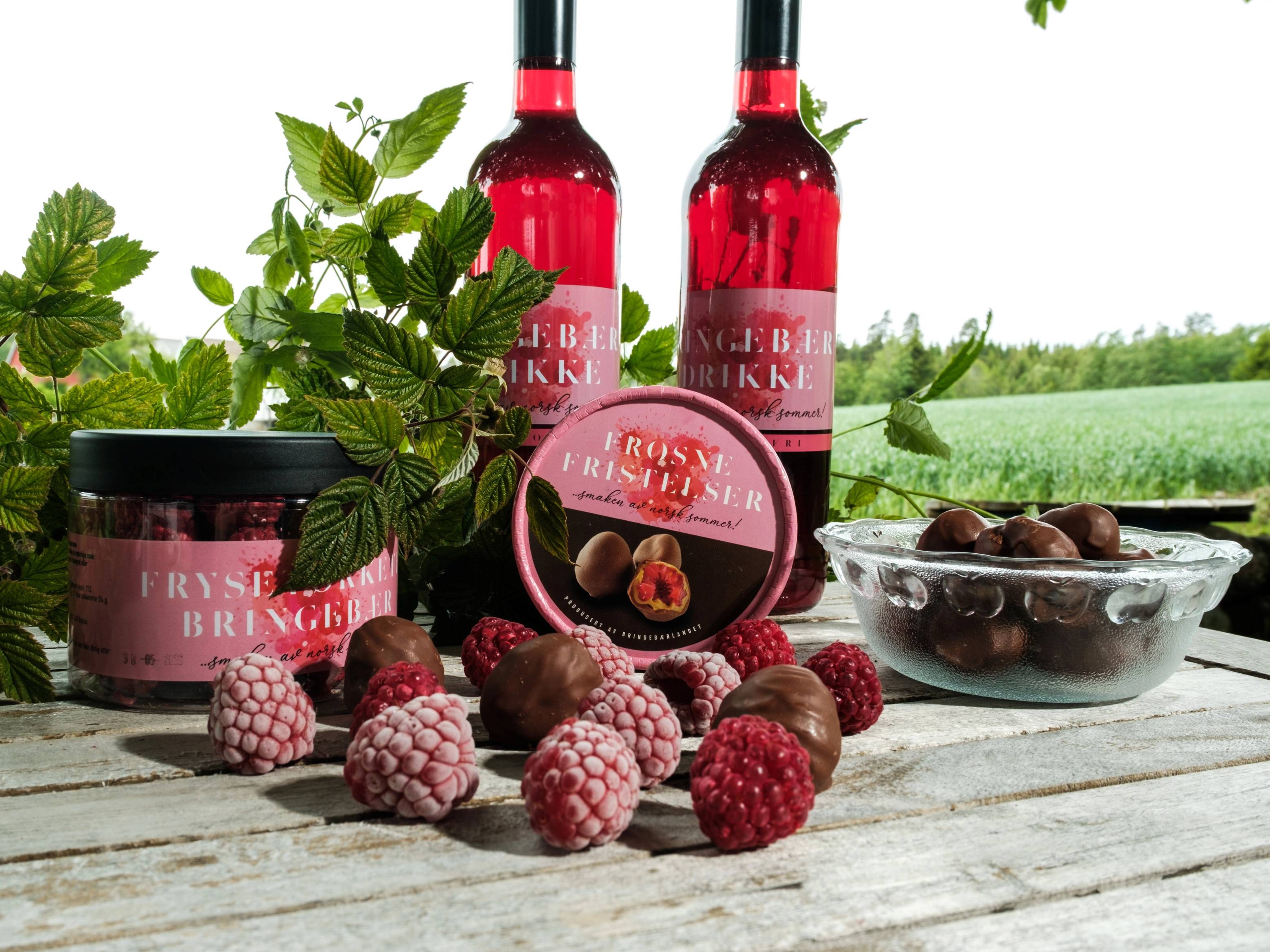 Bringebær, jordbær og bringebærprodukter på et trebord med en åpen mark og trær i bakgrunnen.