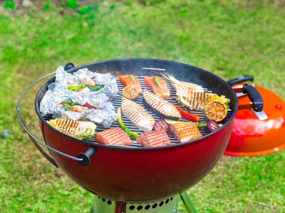 Grill med ulike typer fisk og skalldyr, omgitt av grønnsaker, på en gressplen.