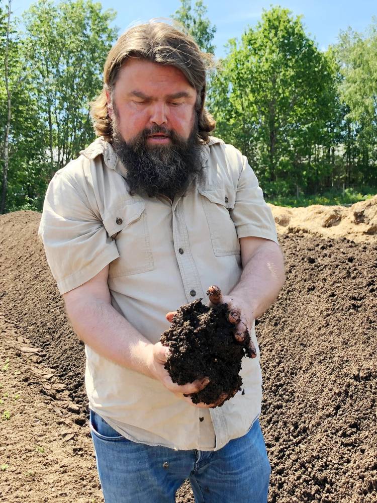 Bjørge Madsen holder en klump med jord i hendene utendørs, med grønt løv og jordbakke i bakgrunnen.