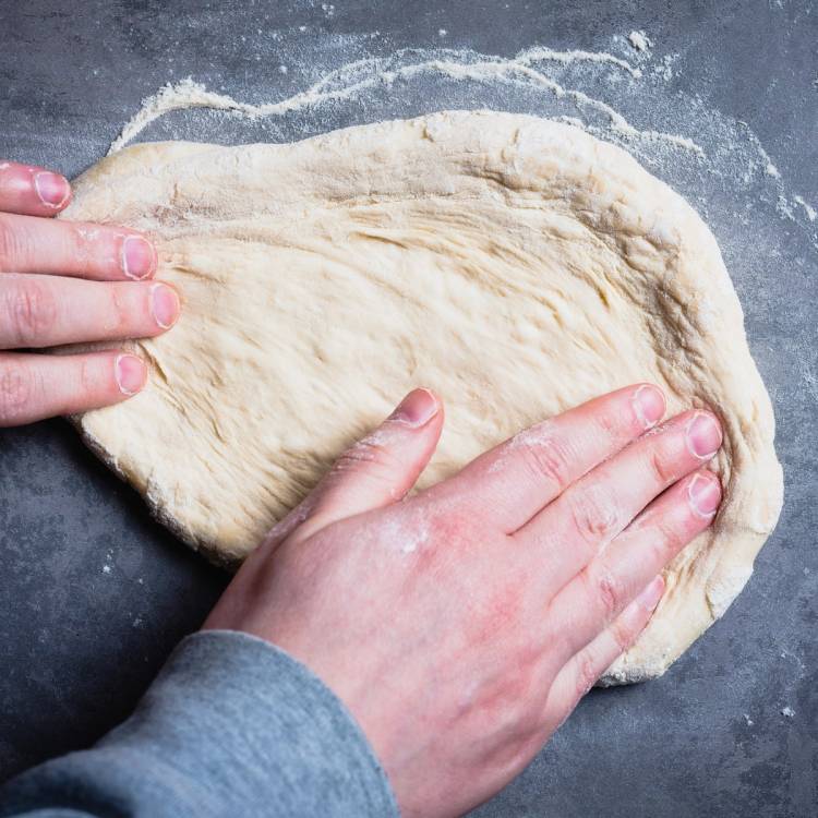 En person former pizzadeig med hendene på en mørk arbeidsflate, med litt mel rundt.