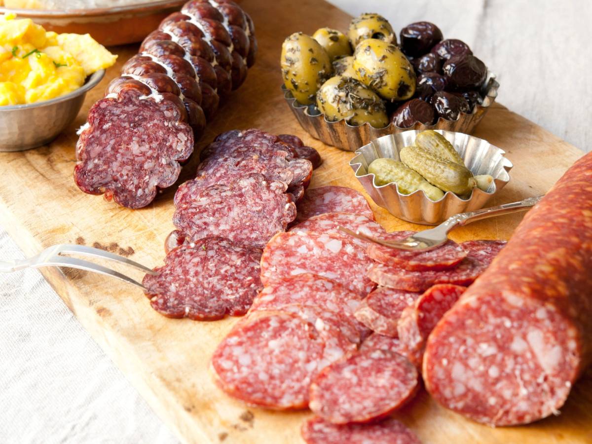 Ulike typer spekemat og pølser på et skjærebrett, med en skål potetsalat og oliven i bakgrunnen.