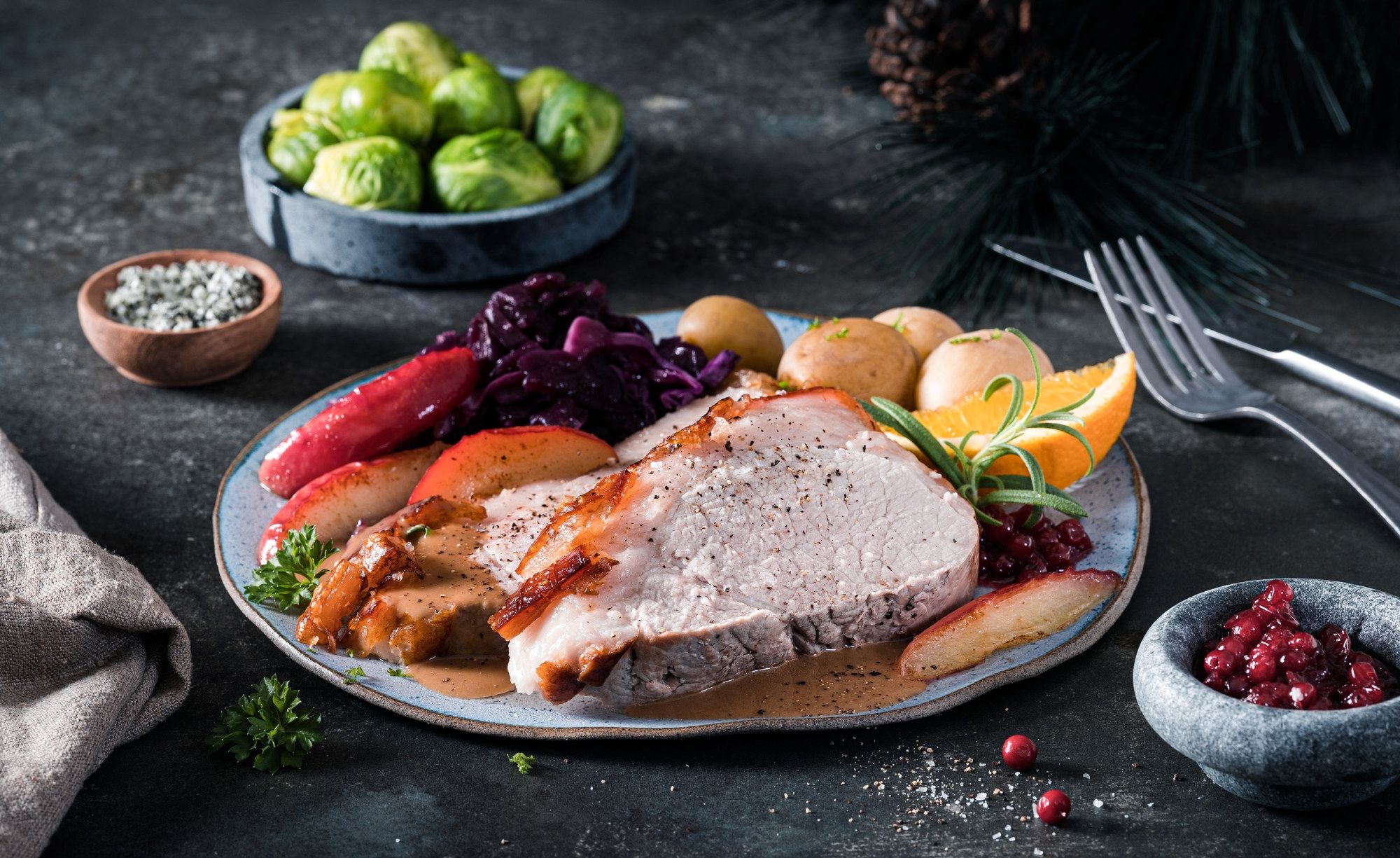 Skinkestek med grønnsaker og tilbehør på en tallerken, omgitt av grønne kål, sitronbåter og tyttebær, på et mørkt bord.