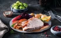 Skinkestek med grønnsaker og tilbehør på en tallerken, omgitt av grønne kål, sitronbåter og tyttebær, på et mørkt bord.