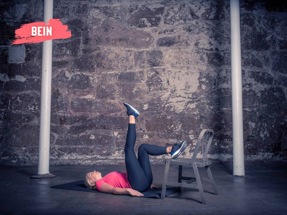 En person ligger på ryggen med beina opp på en stol, utfører et rompeløft-øvelsesposisjon i et treningsrom med murvegg og treningsutstyr i bakgrunnen.